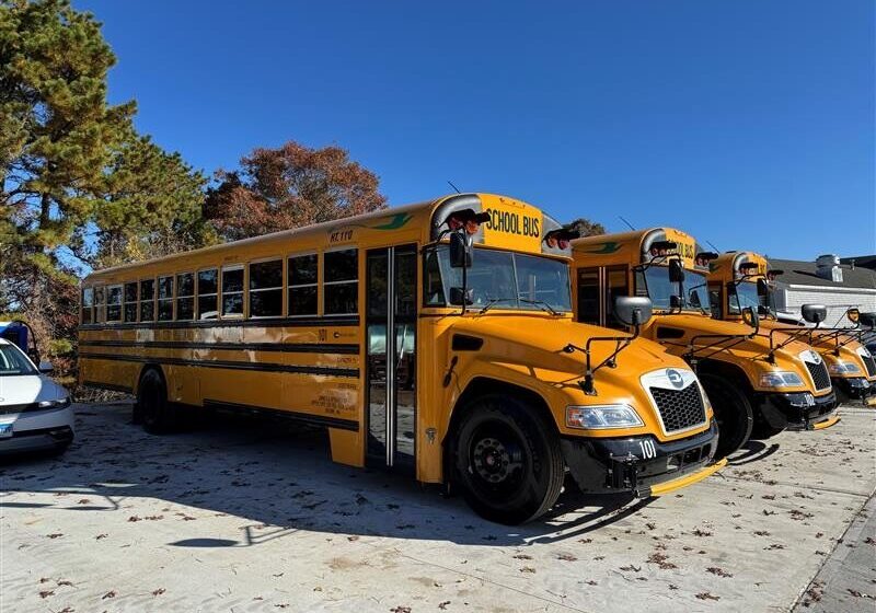 Image 2 - electric school bus webpage Three school buses lined up and parked horizontally. To the left of them is a small white vehicle. Behind them are some autumn colored trees, and some leaves are scattered on the ground around the buses.