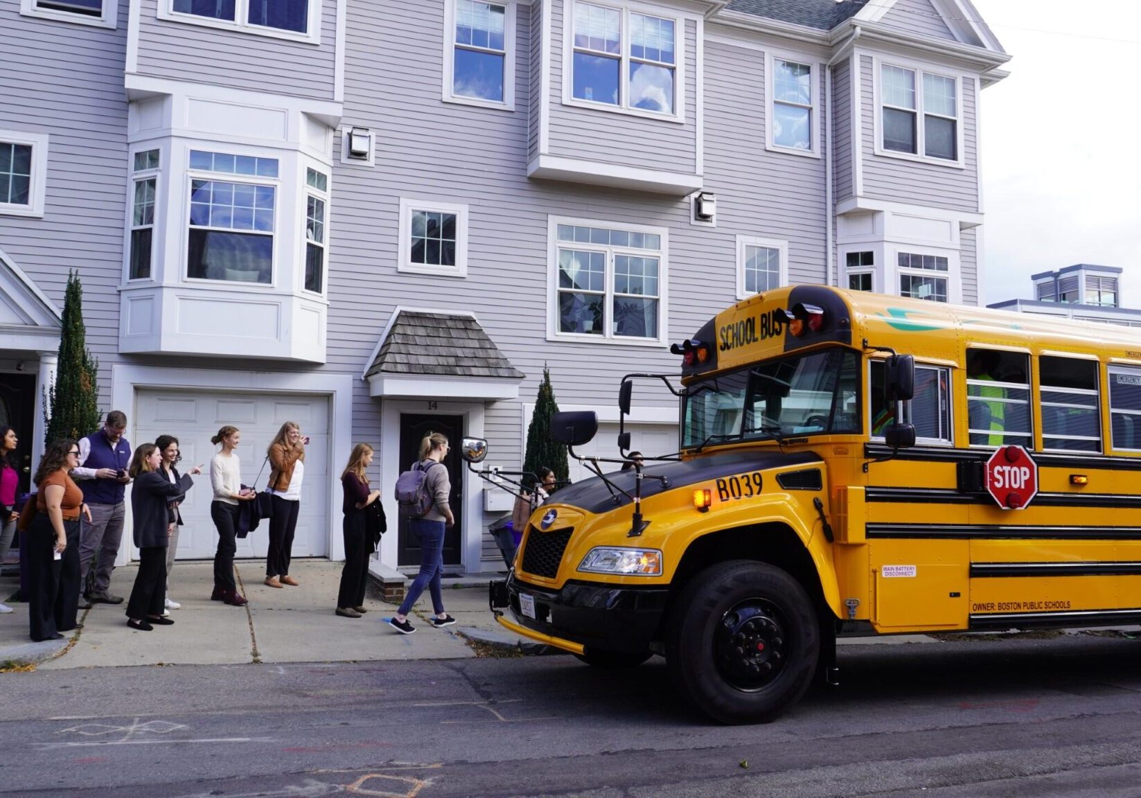 Image 3 - electric school bus webpage MassCEC staff board an electric school bus on the way to attend an infrastructure focused depot tour. They are standing on a sidewalk and there is a large gray apartment or condo building behind them.