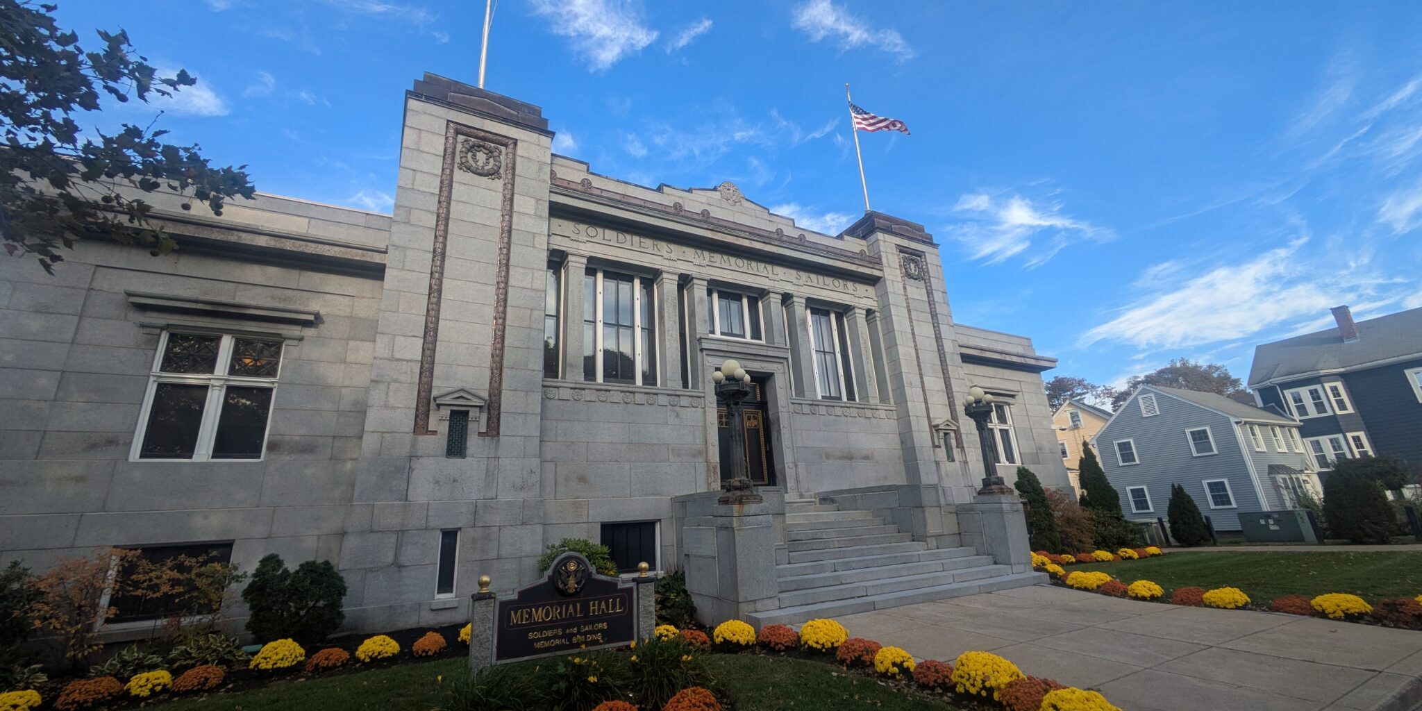 Melrose Memorial Hall from the front. A large building that has steps to the front door, and two American flags on the roof. There are flowers and grass on both sides of the walkway to the front door.