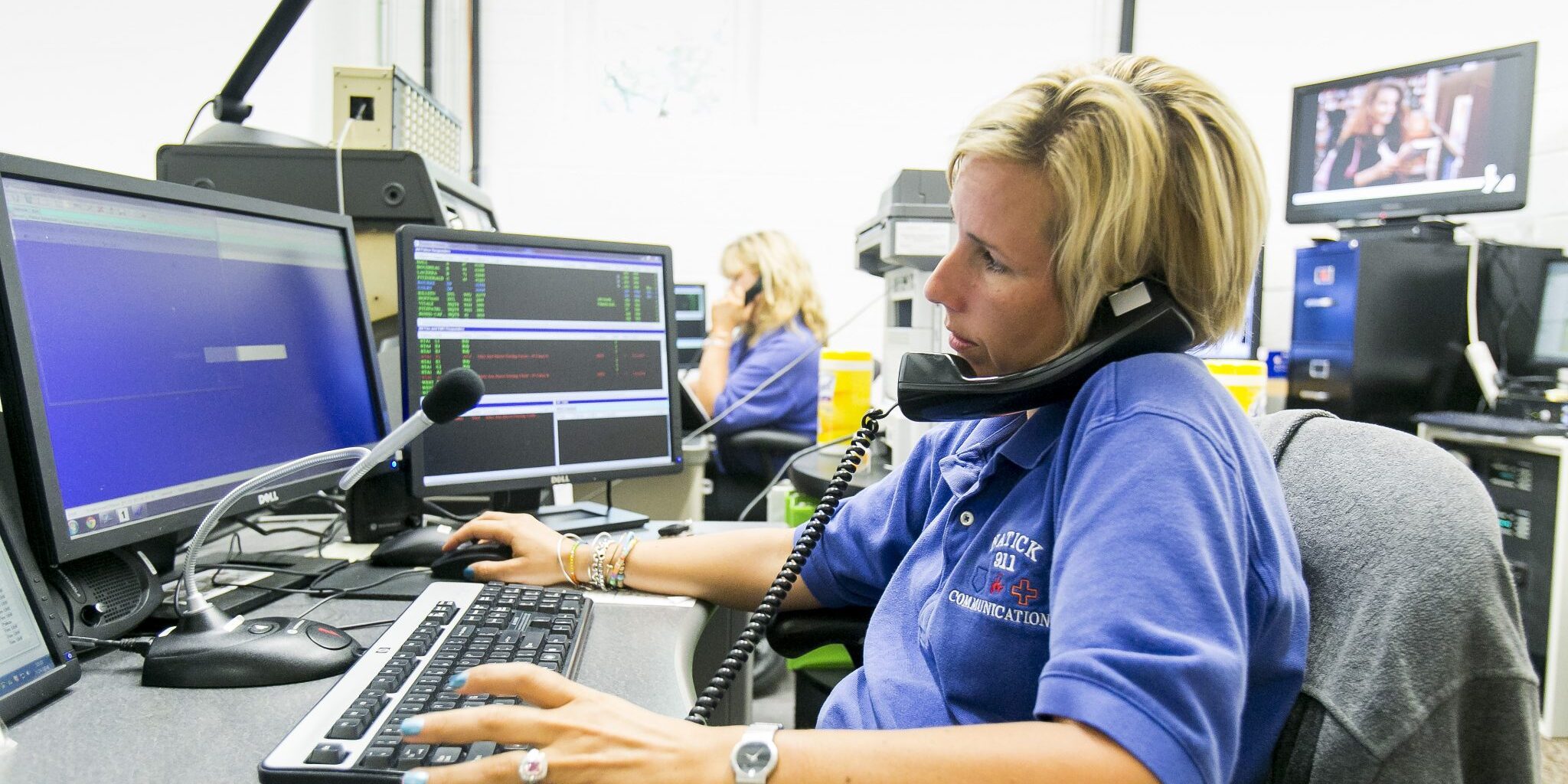 Natick313 Dispatcher Aubrey Colby Photo is of a Natick emergency dispatcher sitting at a computer and she is on the phone talking to someone. She is a Caucasian woman with short, blonde hair, and is wearing a purple colored short-sleeve shirt.