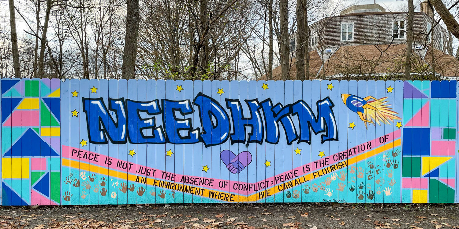 Needham-Mural-1 Image is of a tall fence that has been painted with a mural. The painting has a light purple/blue background. In large blue letters towards the top it says NEEDHAM. Under the word is a painted pink and purple heart made from hands shaking. Towards the bottom is a thin pink line and a yellow line. At the very bottom of the mural it's aqua. Trees are behind the fence and a sidewalk with leaves on it is in front of the fence.