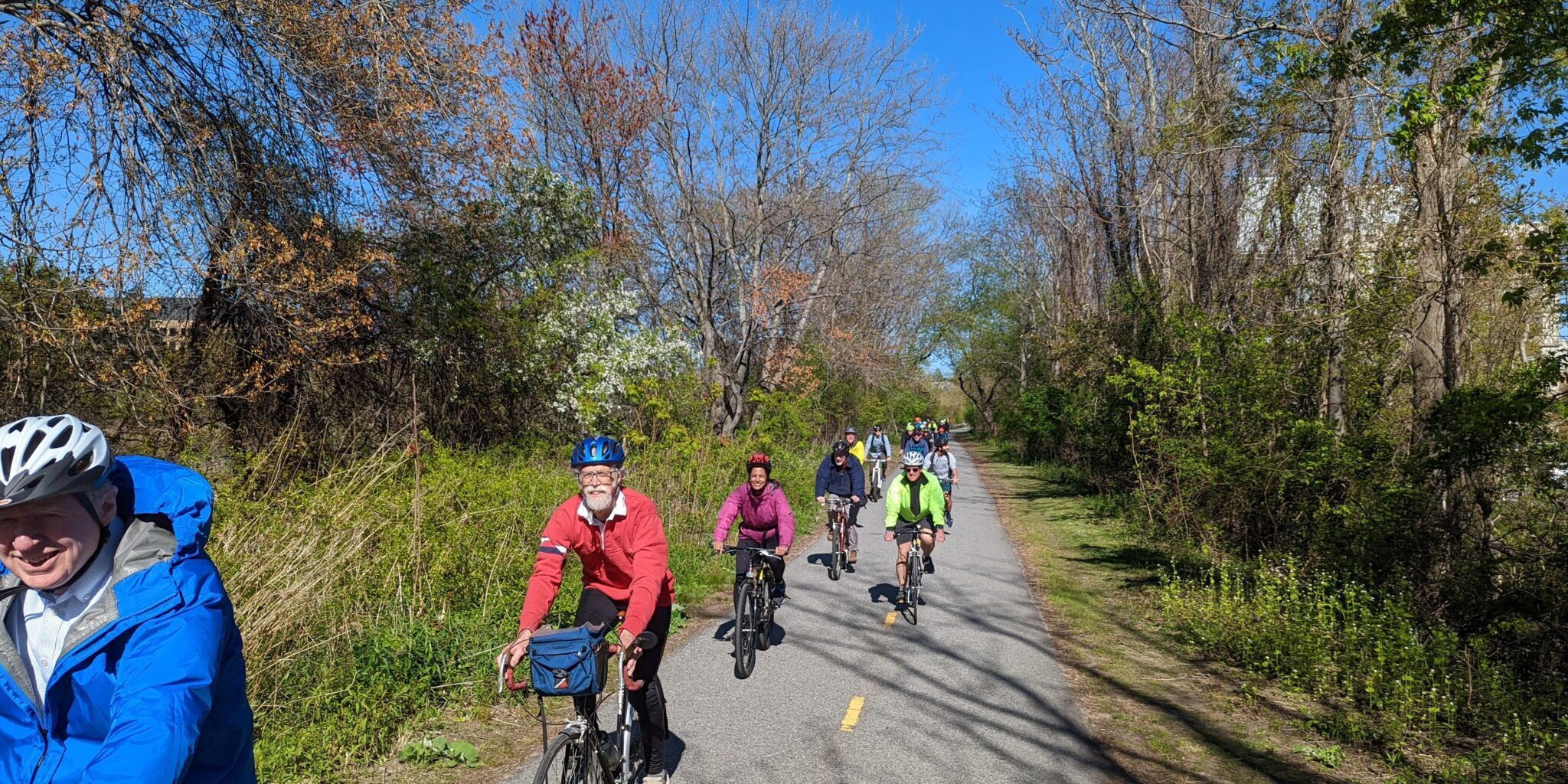 PXL_20220510_201653150 Many people riding bicycles on a concrete road with grass and trees on the sides of the road. It's the Mass Central Rail Trail.
