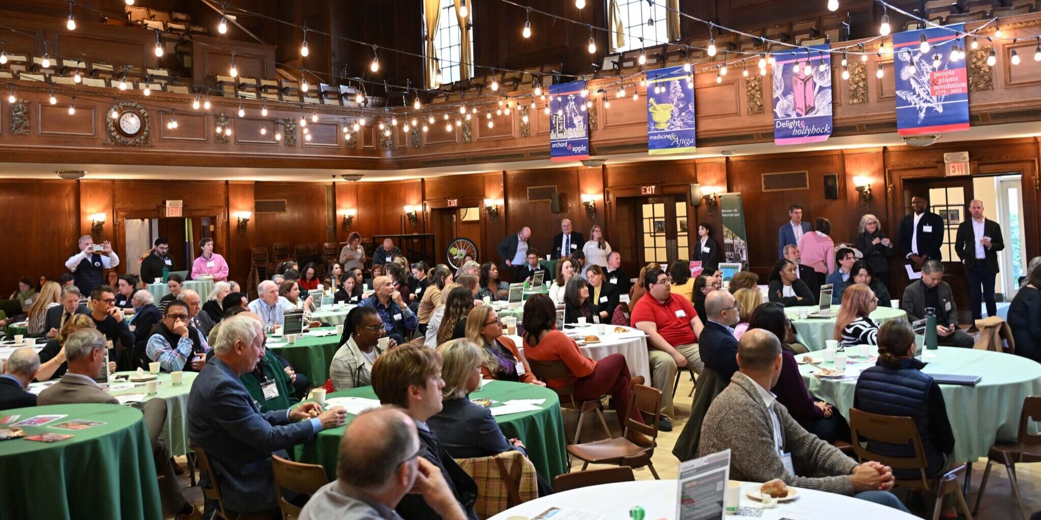 Attendees sit around tables, with most facing away from the camera at MAPC's 2025 Fall Council Meeting. Stringed lightbulbs hang overhead, and an upper gallery of chairs is visible.